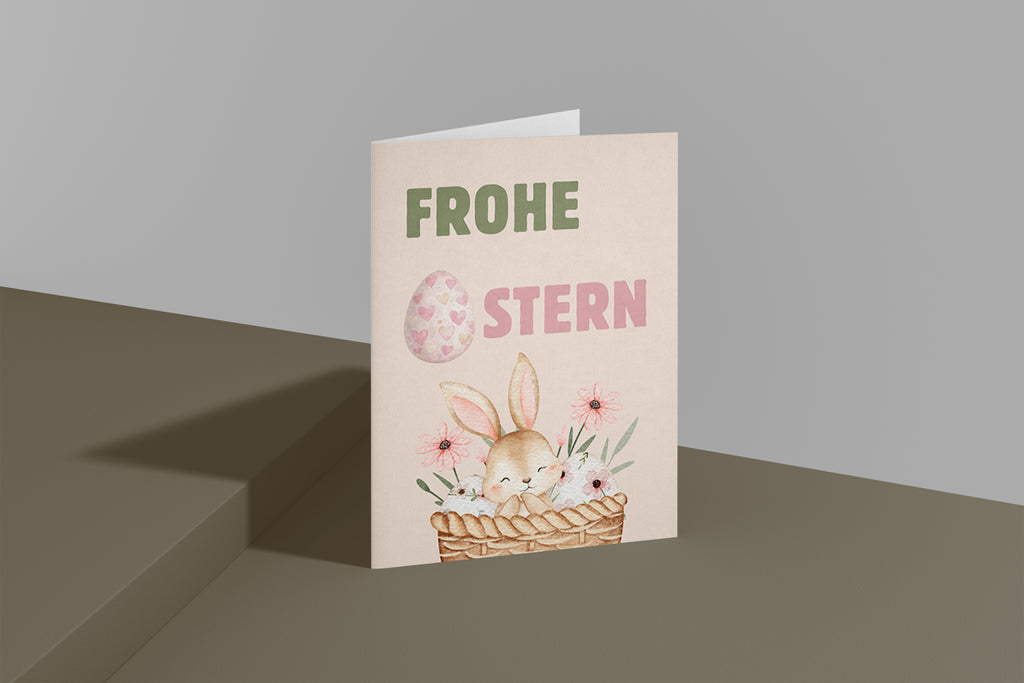 Niedliche Osterkarte mit einem kleinen Hasen in einem Korb mit Blumen und Ostereiern. Aquarell Illustration mit Pastellfarben und dem Schriftzug „Frohe Ostern“. Perfekt als Oster Grußkarte für Familie und Freunde.