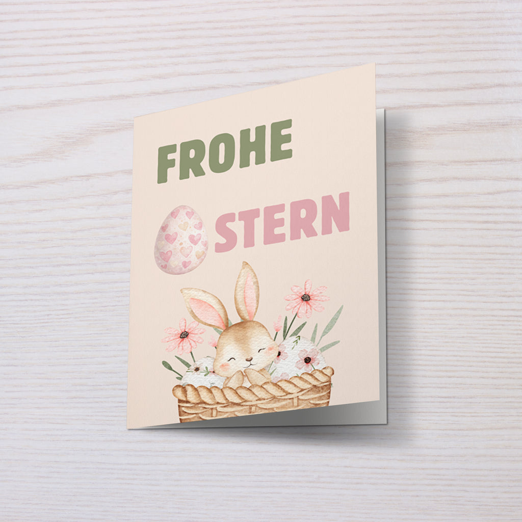 Niedliche Osterkarte mit einem kleinen Hasen in einem Korb mit Blumen und Ostereiern. Aquarell Illustration mit Pastellfarben und dem Schriftzug „Frohe Ostern“. Perfekt als Oster Grußkarte für Familie und Freunde.