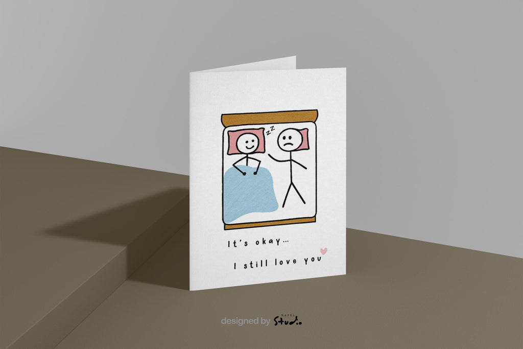 A6-Liebeskarte mit zwei Strichmännchen im Bett: eine Figur schläft lächelnd, die andere liegt wach und wirkt traurig. Darunter steht „It’s okay… I still love you“. Sanfte, humorvolle Illustration über Liebe, Nähe und Beziehungsmomente. Zartes, minimalistisches Design.