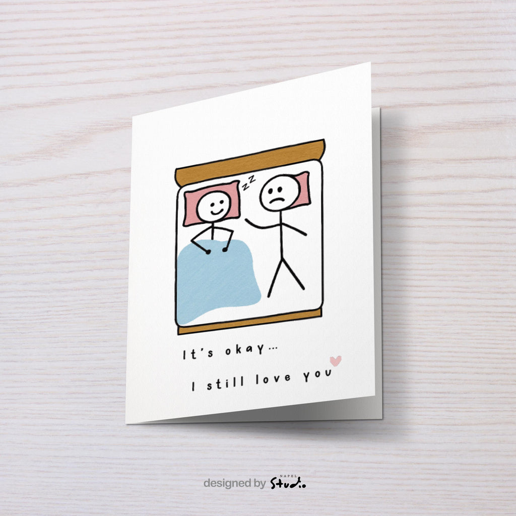 A6-Liebeskarte mit zwei Strichmännchen im Bett: eine Figur schläft lächelnd, die andere liegt wach und wirkt traurig. Darunter steht „It’s okay… I still love you“. Sanfte, humorvolle Illustration über Liebe, Nähe und Beziehungsmomente. Zartes, minimalistisches Design.
