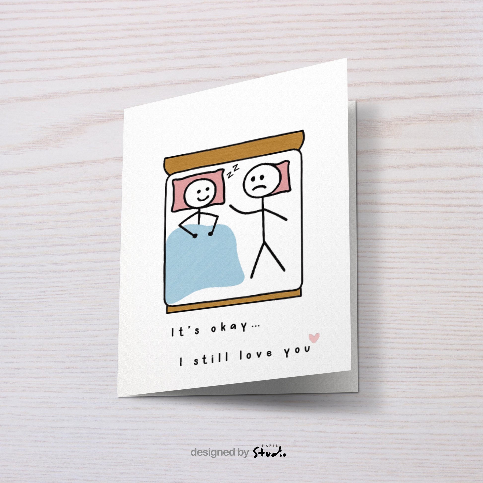 A6-Liebeskarte mit zwei Strichmännchen im Bett: eine Figur schläft lächelnd, die andere liegt wach und wirkt traurig. Darunter steht „It’s okay… I still love you“. Sanfte, humorvolle Illustration über Liebe, Nähe und Beziehungsmomente. Zartes, minimalistisches Design.