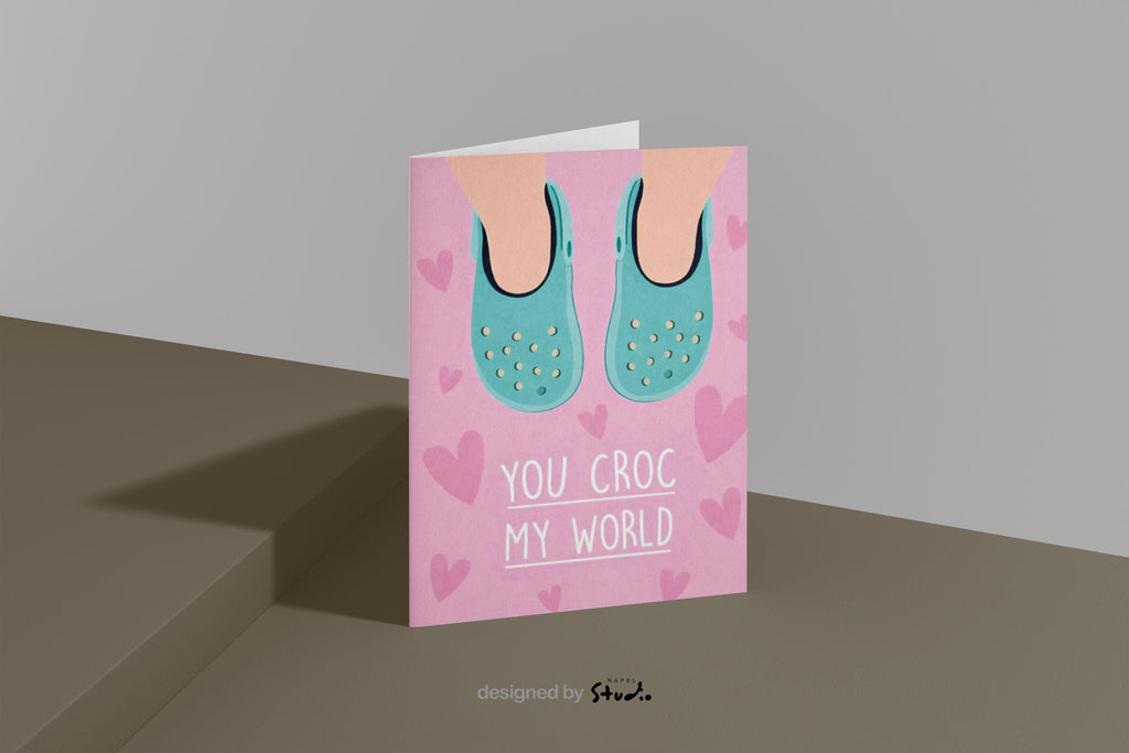 Verspielte Illustration von türkisfarbenen Crocs vor rosafarbenem Hintergrund mit kleinen Herzmotiven. Der Text „You croc my world“ nutzt ein humorvolles Wortspiel und macht die Karte liebevoll und witzig zugleich. Ideal als Valentinstagskarte, Jahrestagskarte oder als lustige Liebeskarte für Partner, Freunde oder Crocs-Fans.