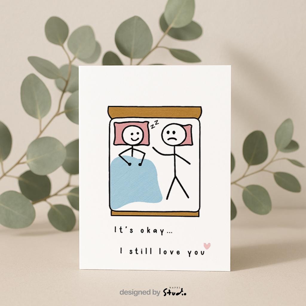 A6-Liebeskarte mit zwei Strichmännchen im Bett: eine Figur schläft lächelnd, die andere liegt wach und wirkt traurig. Darunter steht „It’s okay… I still love you“. Sanfte, humorvolle Illustration über Liebe, Nähe und Beziehungsmomente. Zartes, minimalistisches Design.