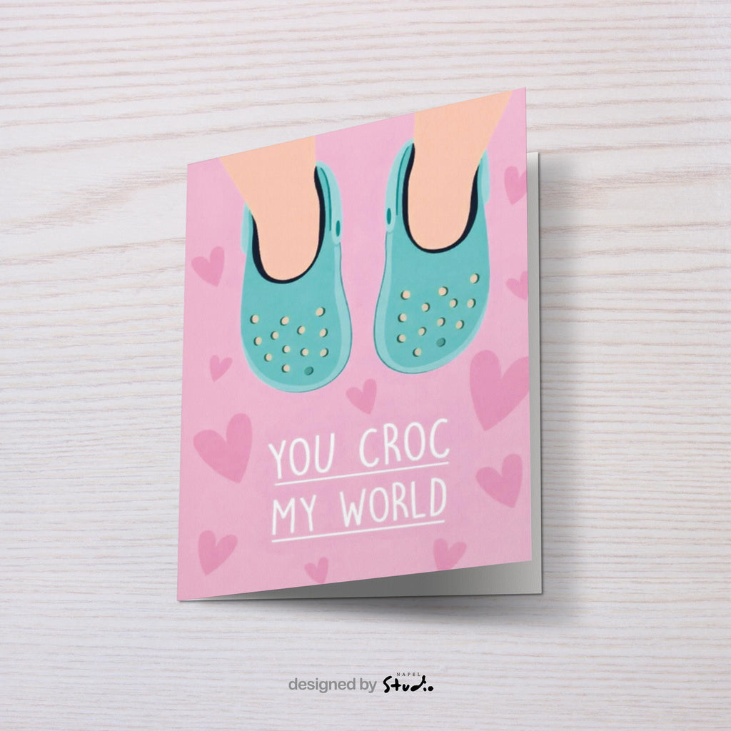 Verspielte Illustration von türkisfarbenen Crocs vor rosafarbenem Hintergrund mit kleinen Herzmotiven. Der Text „You croc my world“ nutzt ein humorvolles Wortspiel und macht die Karte liebevoll und witzig zugleich. Ideal als Valentinstagskarte, Jahrestagskarte oder als lustige Liebeskarte für Partner, Freunde oder Crocs-Fans.
