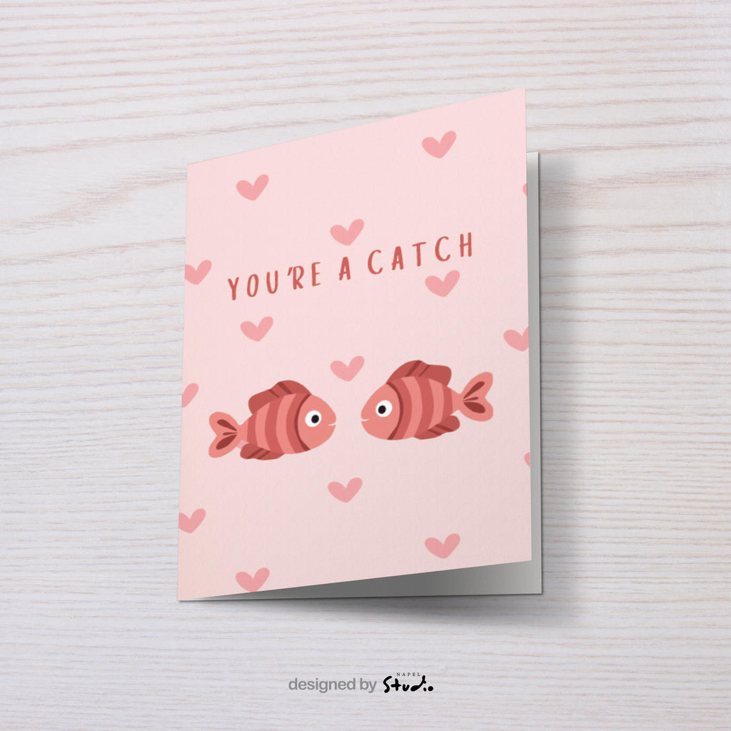 Illustration von zwei Fischen, die sich gegenüberstehen, umgeben von kleinen Herzen. Der Text „You’re a catch“ macht die Karte zu einer charmanten Liebeskarte mit Wortspiel. Ideal für Valentinstag, Jahrestag oder als kleine romantische Aufmerksamkeit.